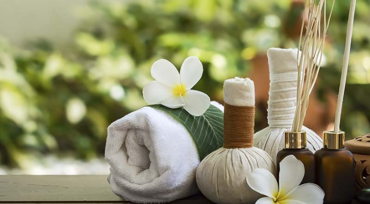 Spa Spa arrangement with a rolled white towel, green leaf, white plumeria flowers, and brown essential oil bottles and diffuser sticks on a wooden surface.