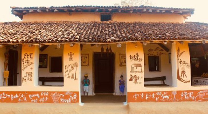 Traditional clay house with a tiled roof and orange mural panels depicting people along the walls. Two children stand in the doorway.