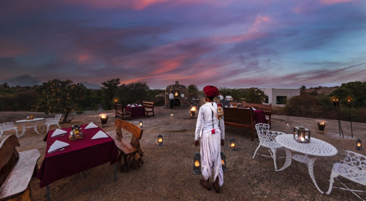 Jungle Dinner Server in white attire and a red turban walks toward a stone fireplace in an outdoor, lantern-lit dinner setup at sunset.