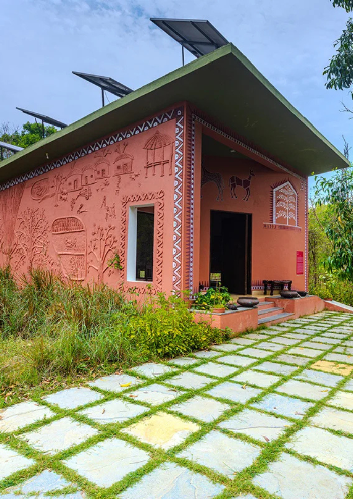 Reddish-orange building with carved wall murals and camel motifs, a dark doorway, and a green sloped roof with solar panels, set beside a grassy, stone-grid path.
