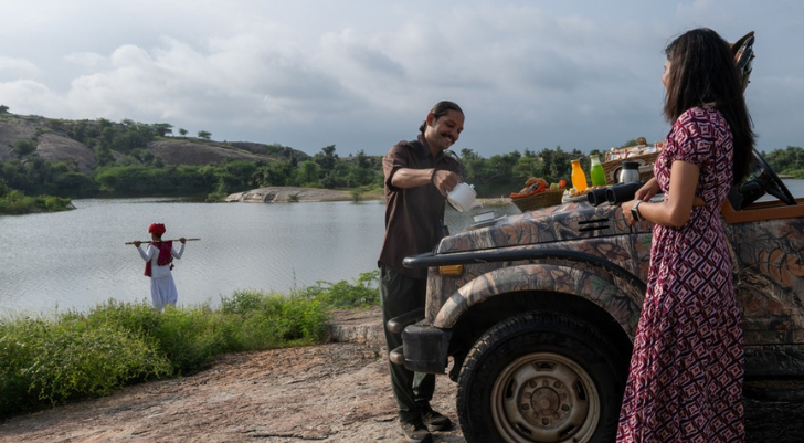 Lakeside Breakfast Man pours a drink from a white jug into a cup beside a camouflaged SUV as a woman in a patterned dress watches nearby by a lakeside road.