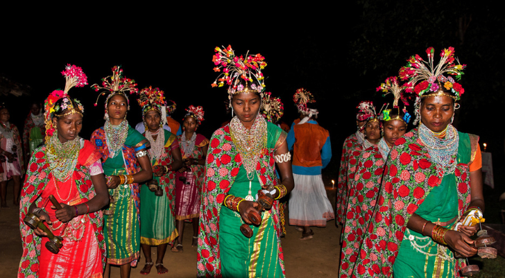 Group of women in bright traditional dresses with floral headpieces walking at night in a cultural procession, holding small wooden items.