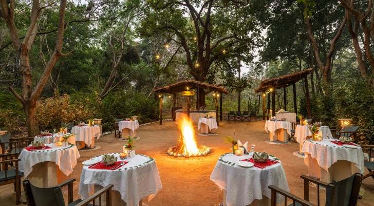 Outdoor dining area around a central fire pit; round tables with white linens, red napkins, and simple centerpieces among trees.