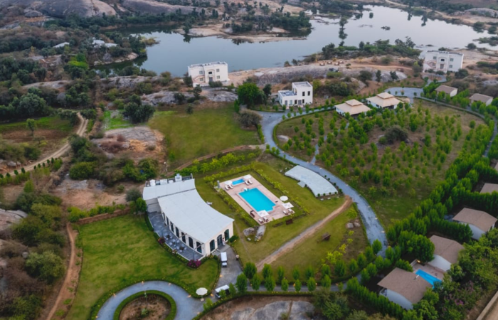 Aerial view of a villa complex with a blue rectangular pool, circular driveway, and manicured lawns surrounded by trees.