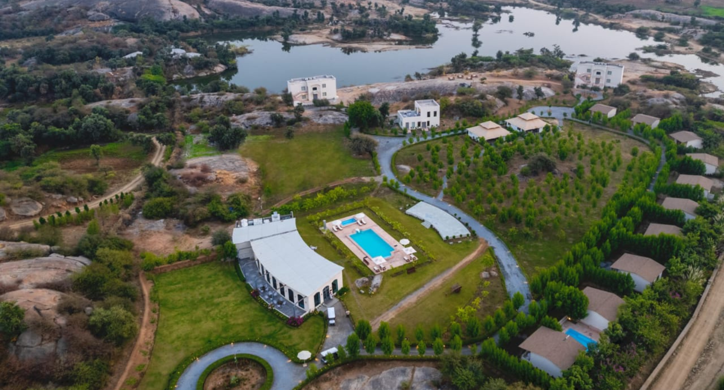 Aerial view of a villa complex with a blue rectangular pool, circular driveway, and manicured lawns surrounded by trees.