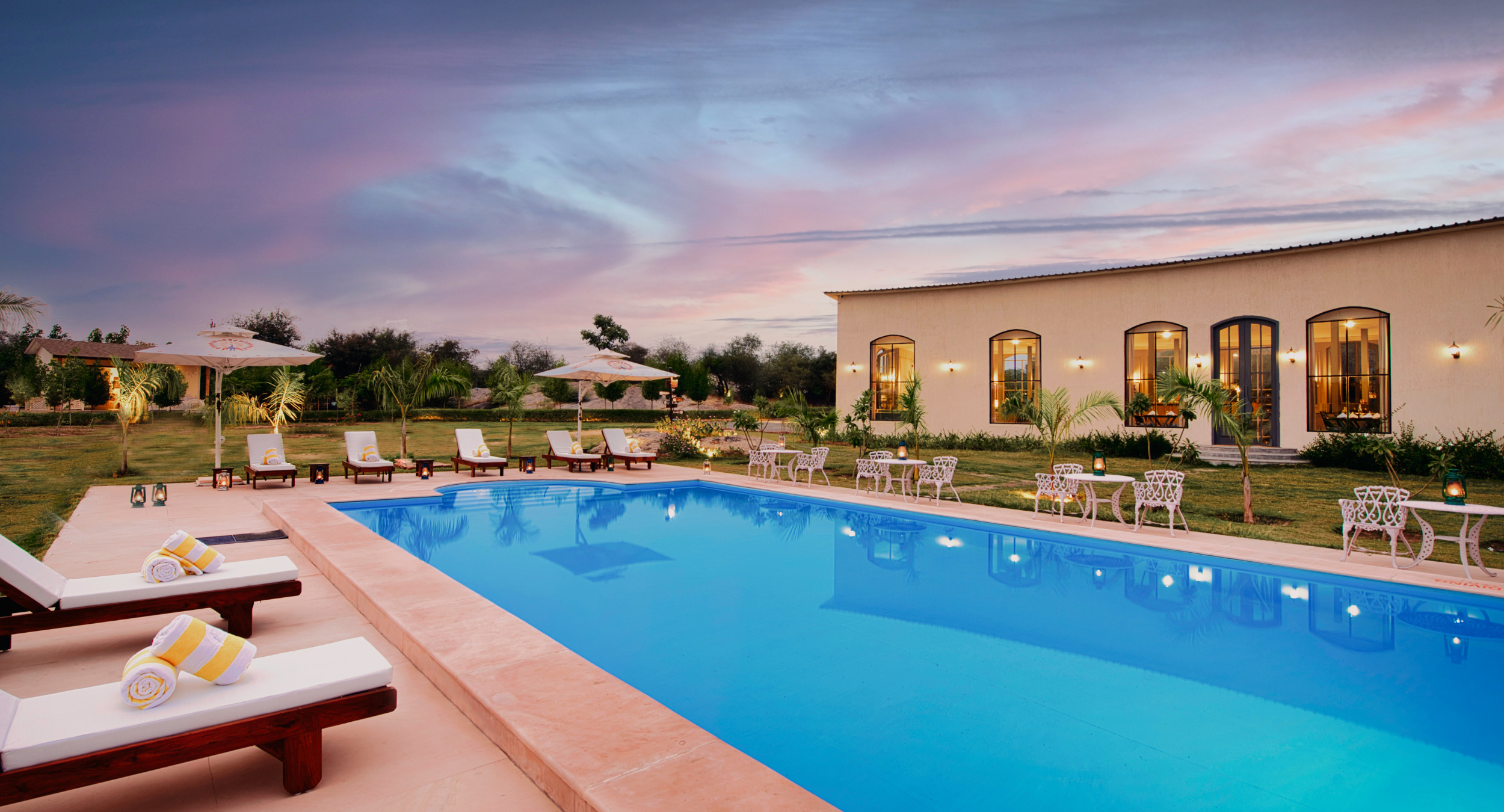 Luxurious resort pool at sunset with white lounge chairs, umbrellas, and a beige building with arched windows.