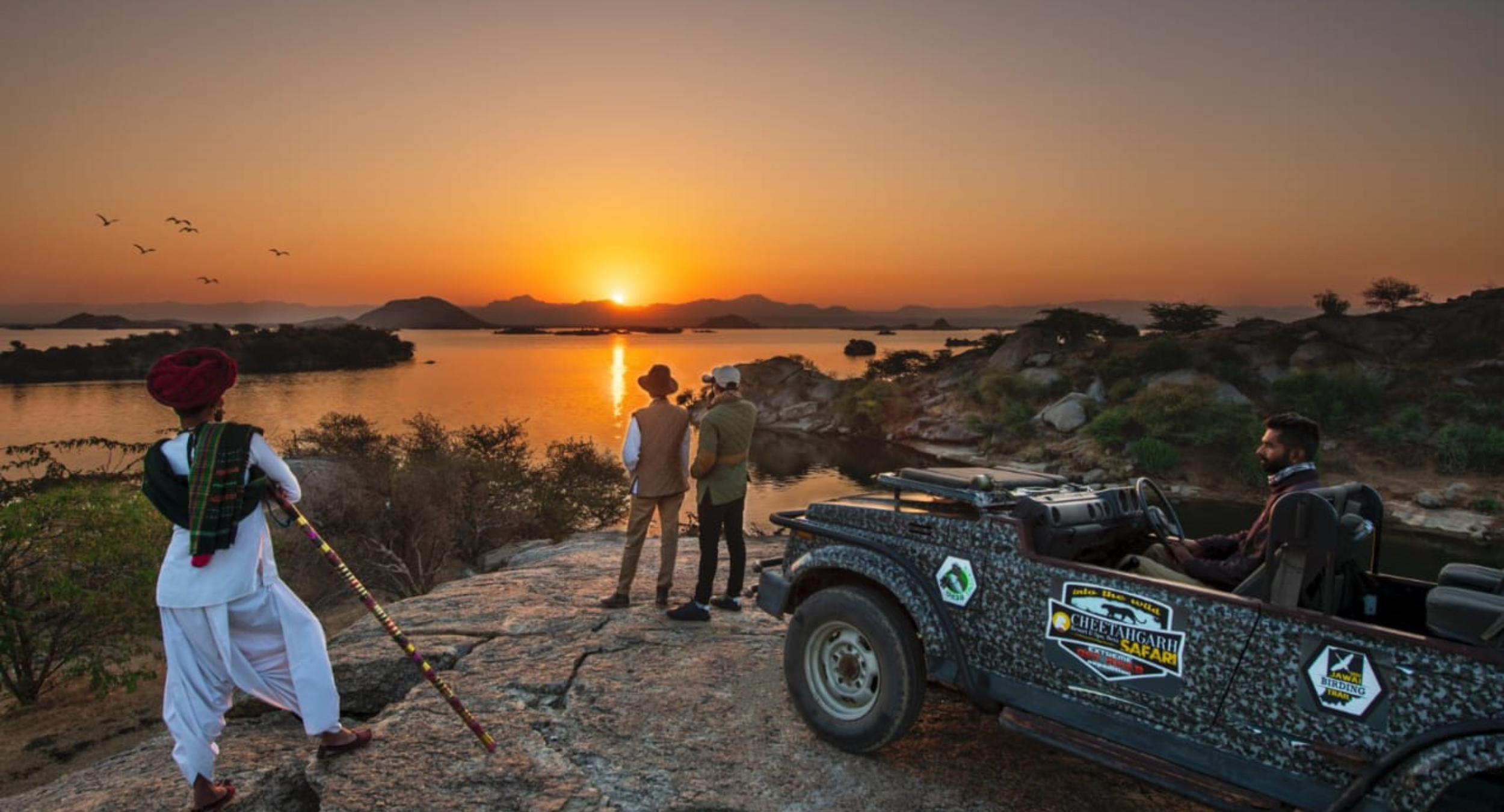 Sunset over a lake with rocky shore as three people in safari outfits stand by a camouflaged jeep, watching the horizon.