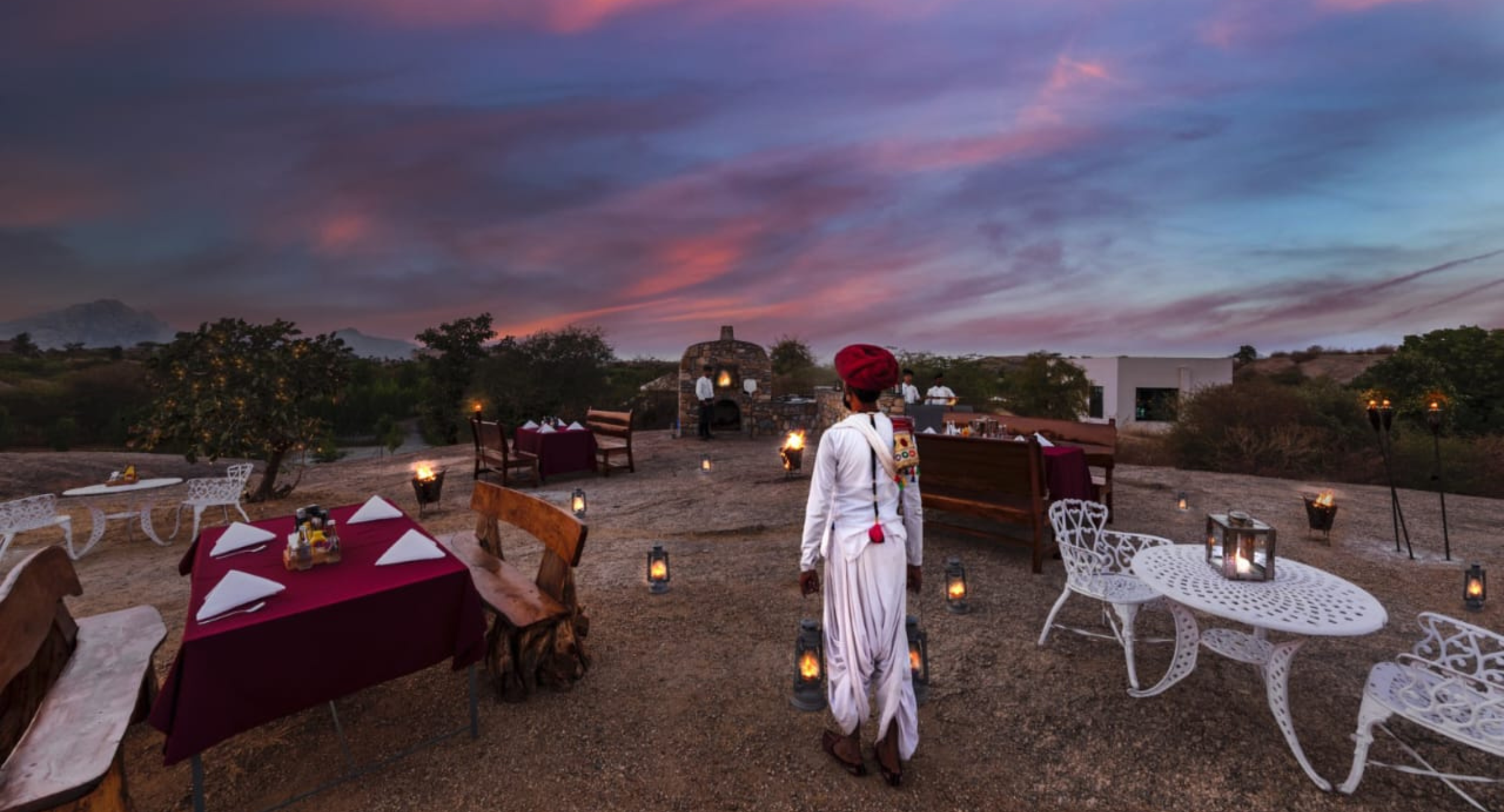Evening outdoor dining on a rocky patio at sunset, with burgundy-cloth tables, lanterns, white iron chairs, and a server in white robes and a red turban by a stone oven.