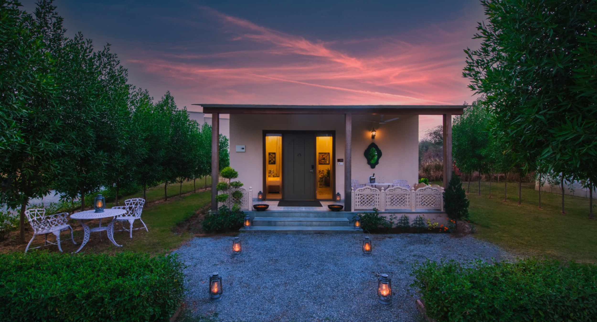 Cozy single-story home with a covered porch, warm lights in the doorways, white wrought-iron table and chairs, and lanterns along a gravel path at sunset.