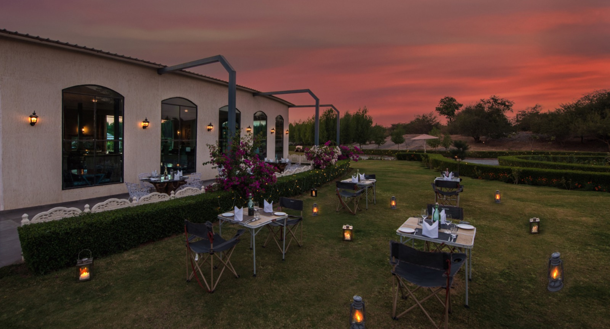 Outdoor restaurant dining area at sunset with tables set and lanterns along the lawn. Tents? No, open-air tables beside a light-colored building.