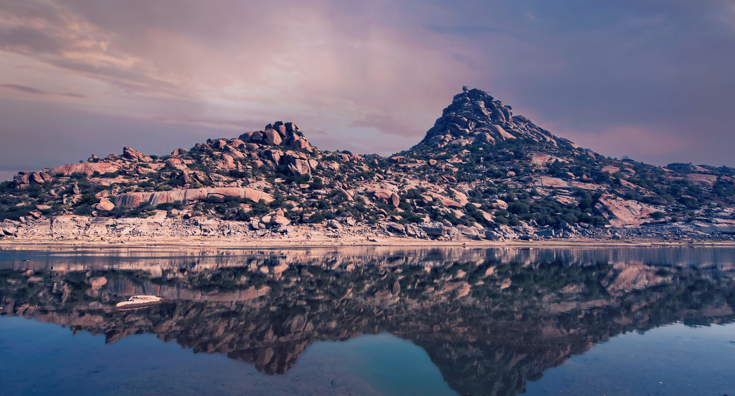 Rocky hill landscape with a prominent peak, reflected in calm water at sunset.