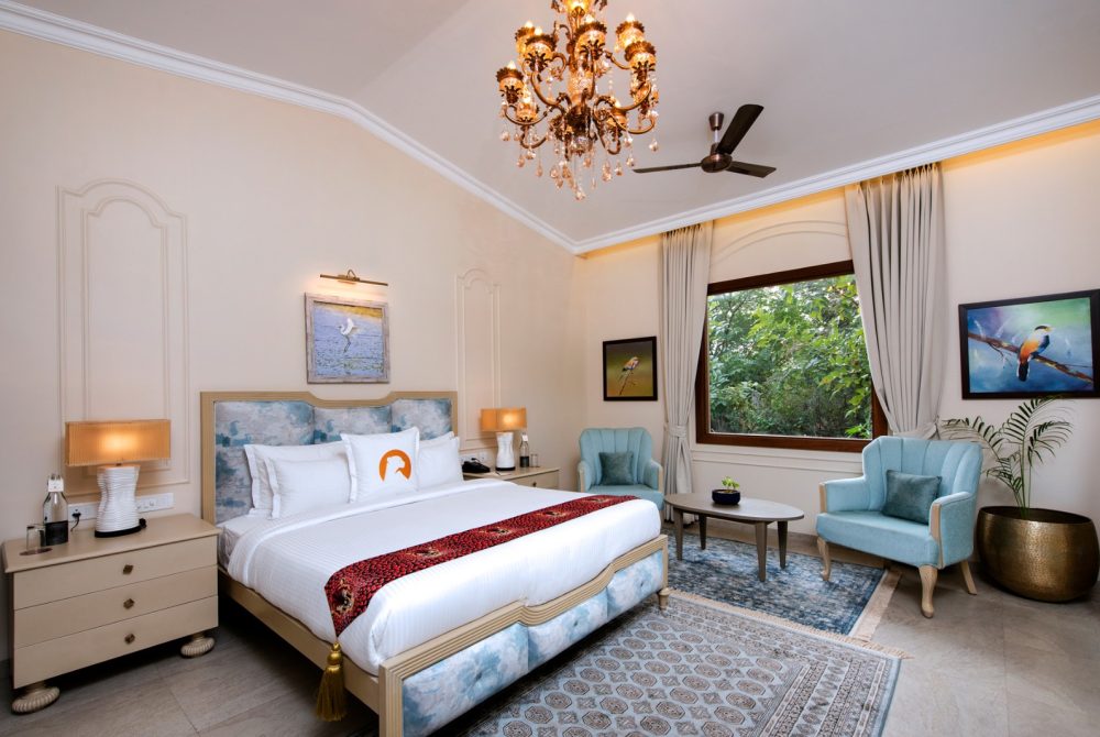 Luxurious hotel bedroom with a crystal chandelier, king bed with blue upholstered headboard, and a seating area by a large window.