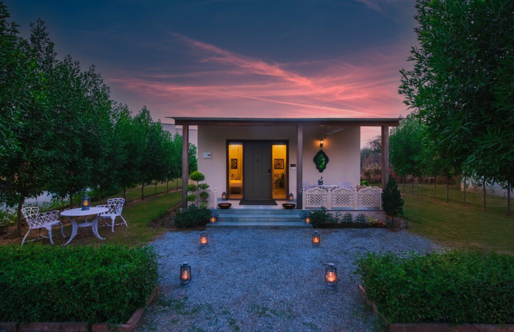 Cozy cottage with a warmly lit front door, white wrought-iron seating on the porch, and lanterns along a gravel path at sunset.