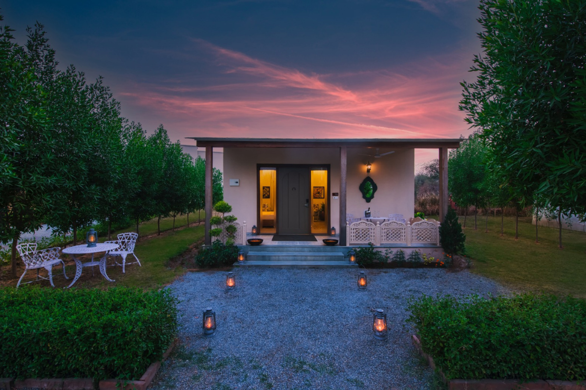 Cozy single-story house with a covered porch, warm lights, and a gravel path lined with lanterns leading to the front steps at dusk with trees around.