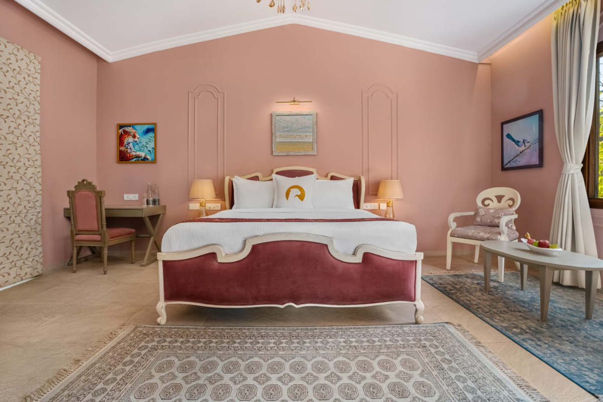Elegant pink hotel bedroom with a grand upholstered bed, matching chairs, and a seating area by a window.