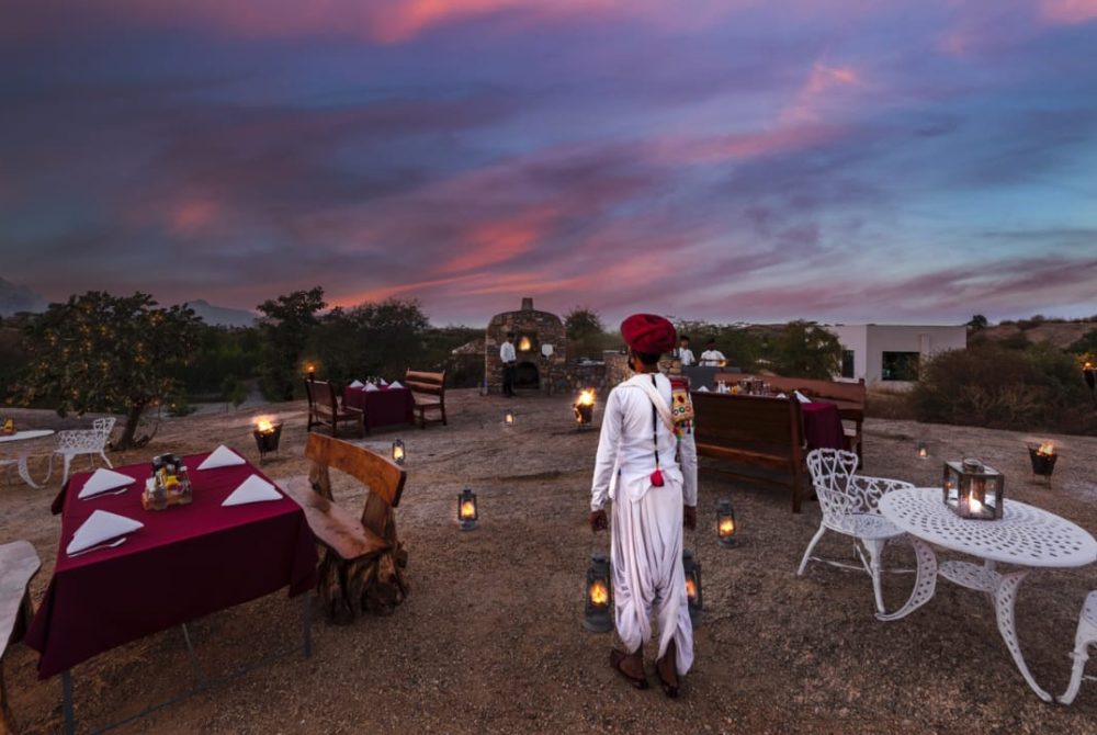 Outdoor evening dining setup in a desert landscape with burgundy tablecloths, white wrought-iron furniture, and lanterns, under a colorful sunset sky.