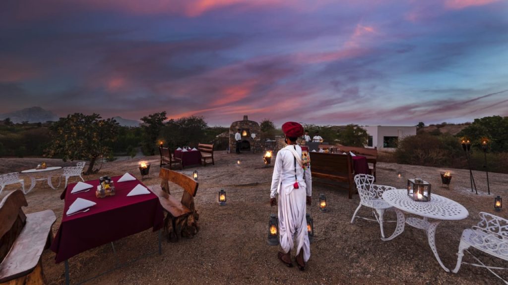 Outdoor evening dining setup in a desert landscape with burgundy tablecloths, white wrought-iron furniture, and lanterns, under a colorful sunset sky.