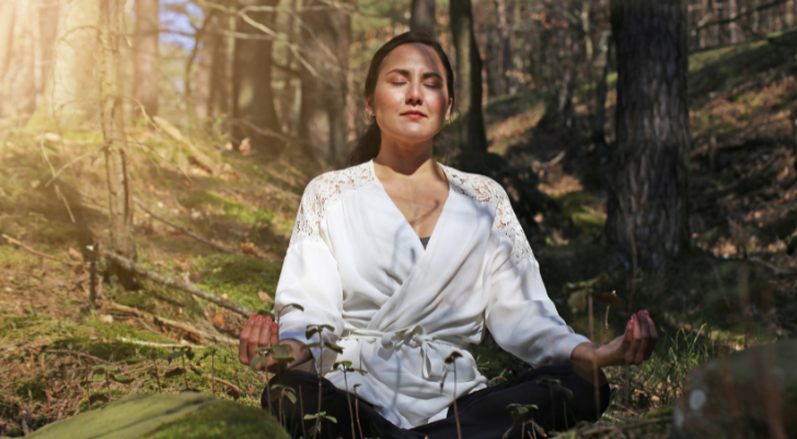Woman sits cross-legged in a forest clearing, eyes closed, wearing a white robe and meditating.