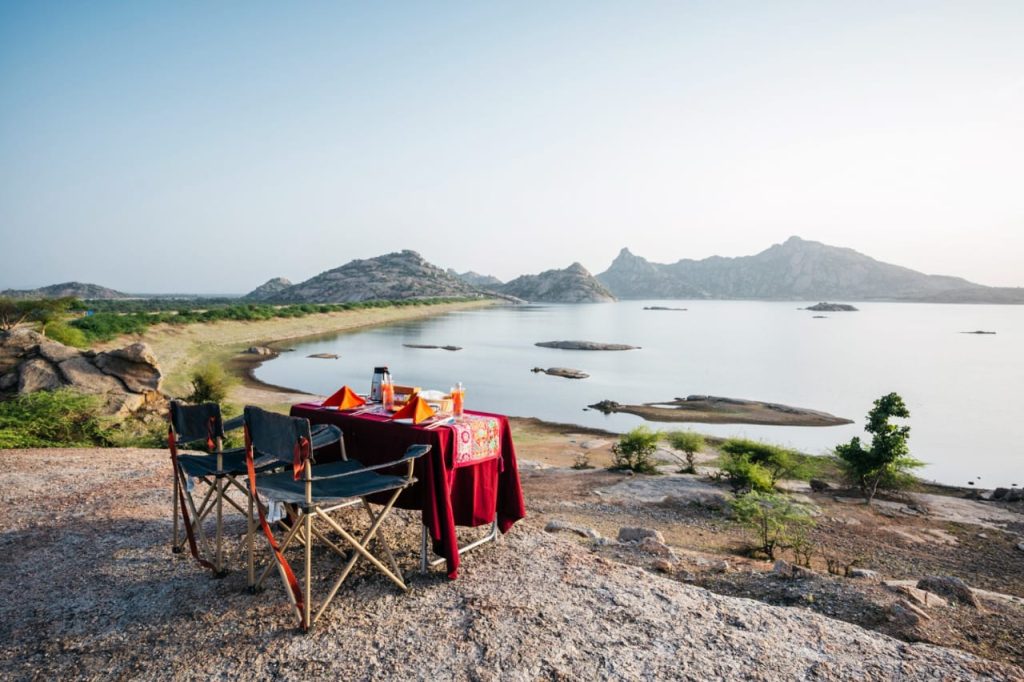 Romantic outdoor lakeside dinner setup with a red tablecloth, two chairs, and place settings by a rocky shore and calm water.