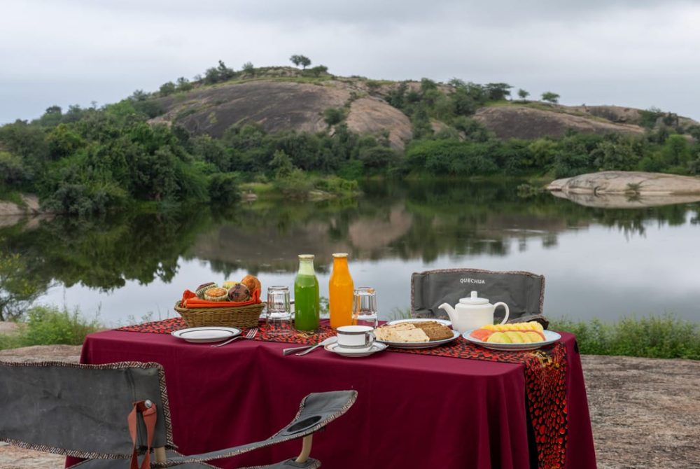 Picnic setup on a red tablecloth by a calm lake with hills, bread, juice bottles, teapot, and plates visible.