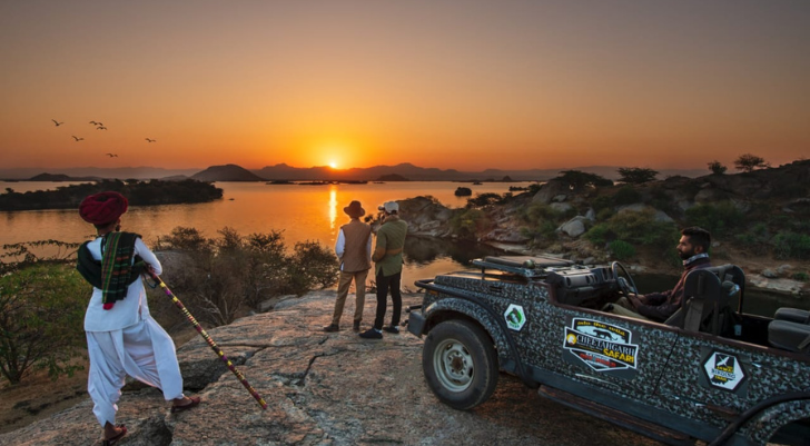 Sunset over a calm river at a rocky overlook; a man in traditional dress holding a staff on the left, two people standing near center, a camo-patterned jeep on the right.