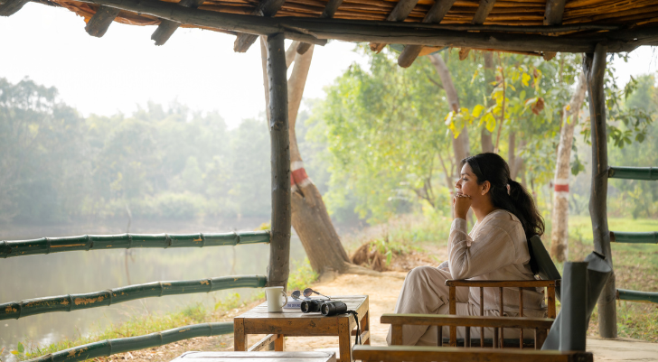 Seated woman in white, under a thatched shelter by a lake, with a camera and mug on a small table nearby.