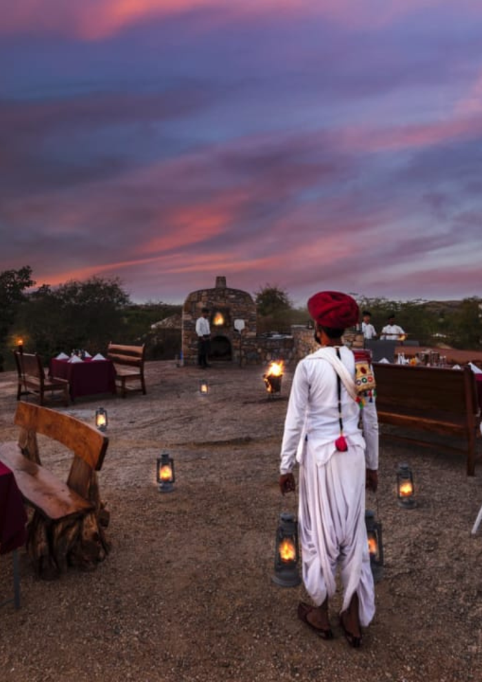 JUNGLE DINNER A person in white traditional attire and a red turban stands among lanterns at an outdoor rustic dining setup at sunset.
