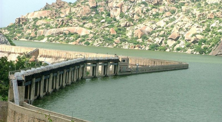 Curved concrete dam extending into a greenish reservoir with rocky hills in the background.