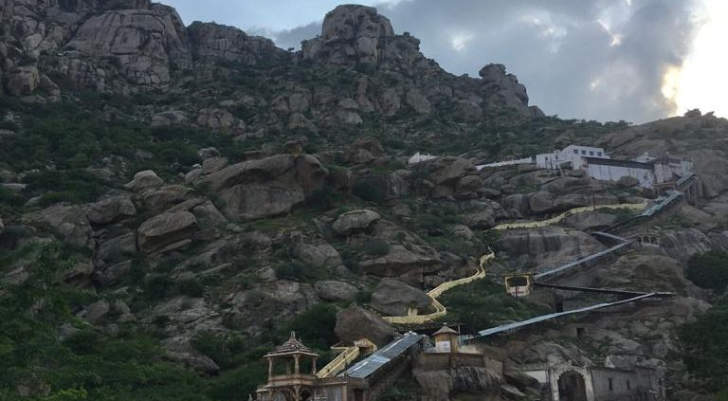 Rocky hillside with a winding yellow-lit path and white buildings climbing toward a hilltop structure, likely a temple complex.