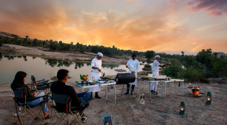 Outdoor riverbank dinner with a barbecue setup at sunset; three chefs cook while two guests sit nearby.