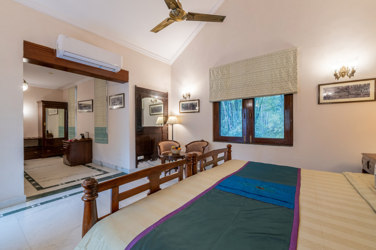 Hotel bedroom with a wooden bed, seating area, dresser, and a window with a view of greenery in soft beige tones.