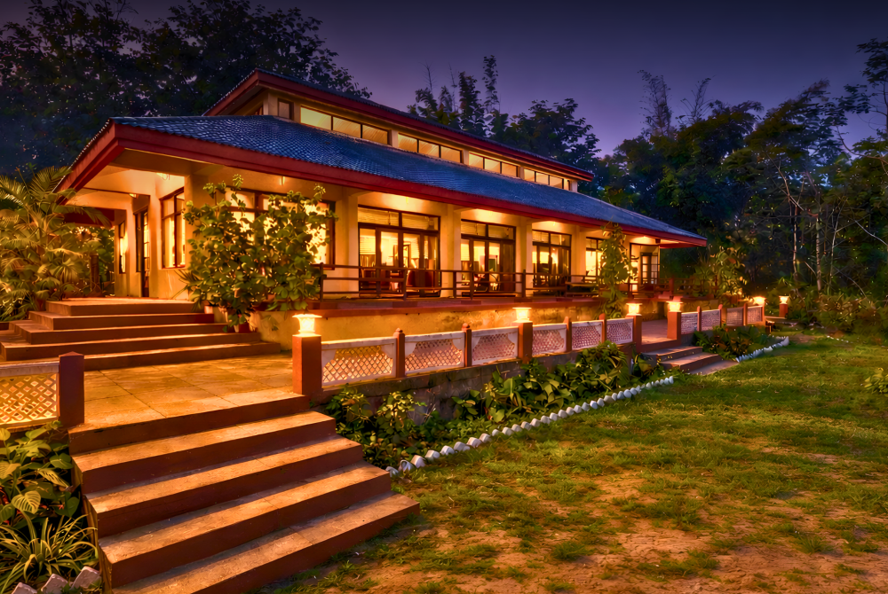 Warm, single-story house with a red roof and glowing windows, set among lush tropical plants and stepped stone stairs at dusk.