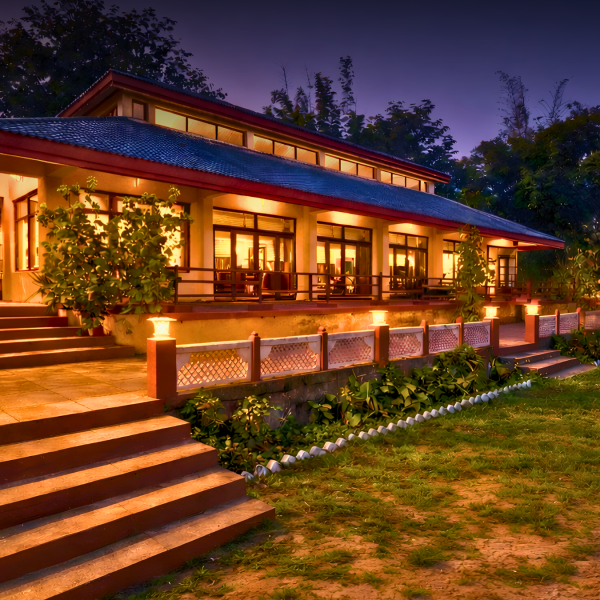 Night view of a warmly lit bungalow with large glass doors, wooden steps, and tropical garden at dusk.