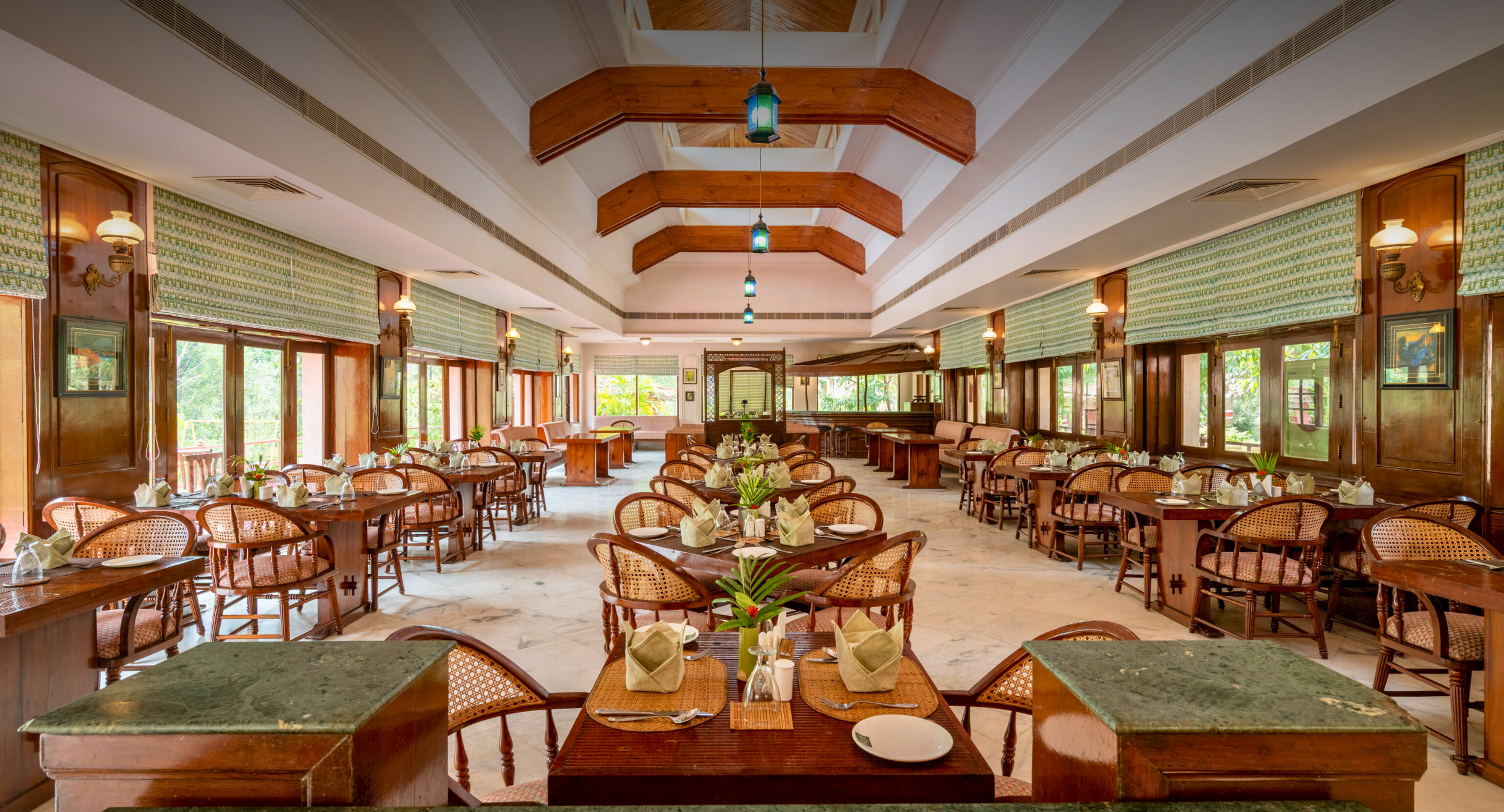 Spacious restaurant dining hall with wooden tables, wicker chairs, green window blinds, and hanging lanterns.