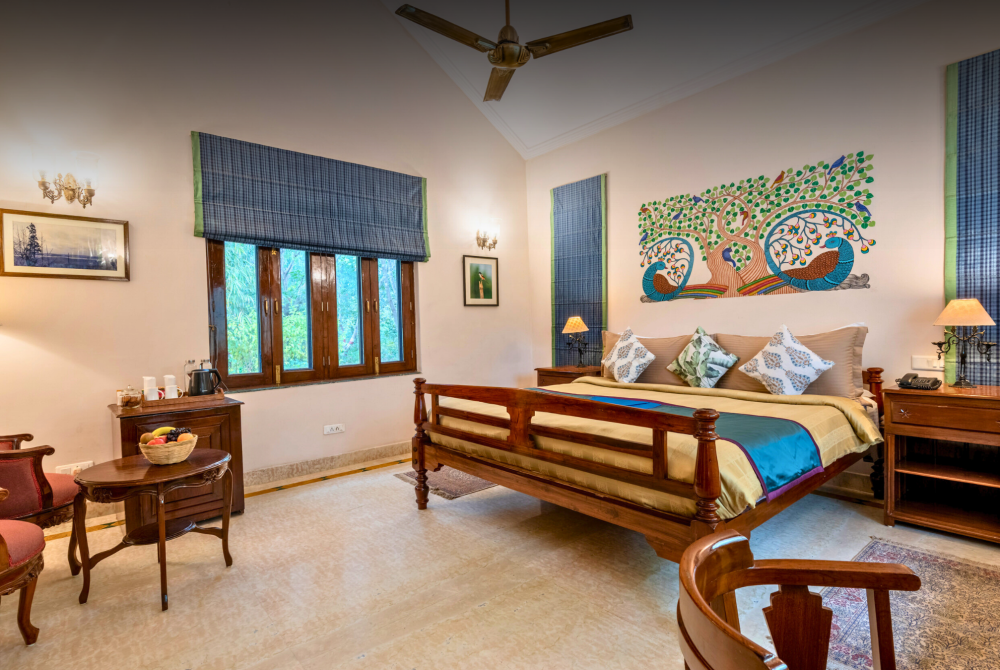 Hotel bedroom with a carved wooden bed, beige bedding, and a vibrant tree-and-peacock mural on the wall above the headboard.