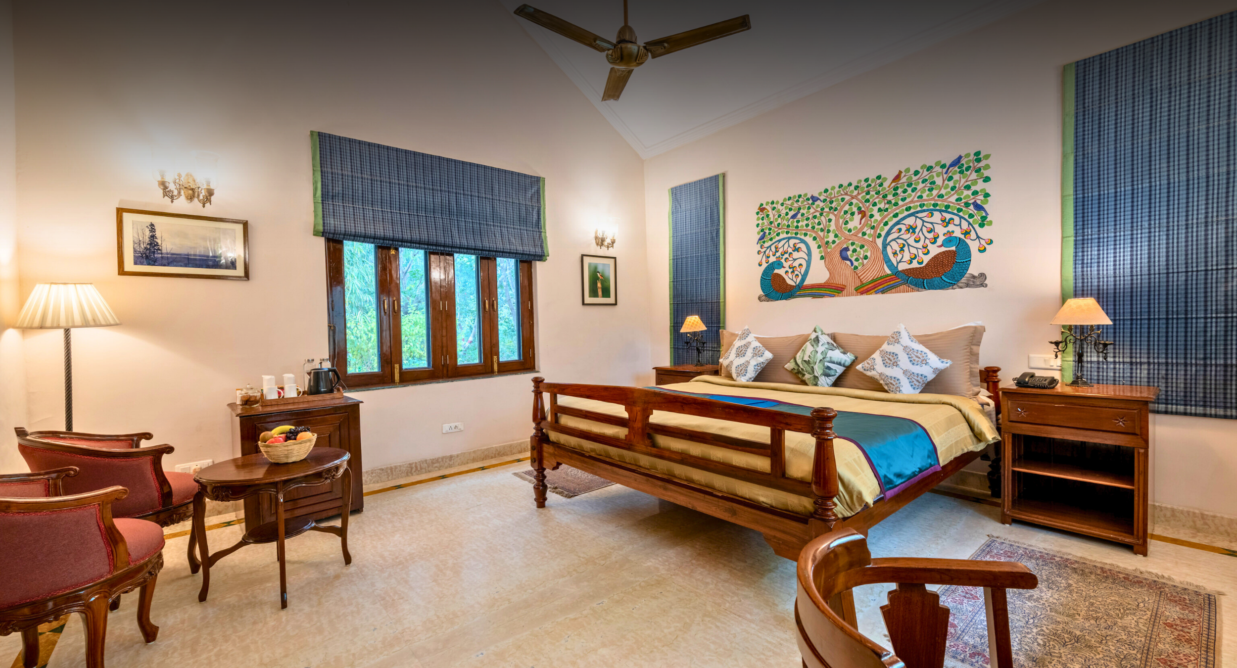 A cozy hotel bedroom with a wooden four-poster bed, colorful bedding, a mural of a tree with peacocks on the wall, and blue-check curtains by a window wall.