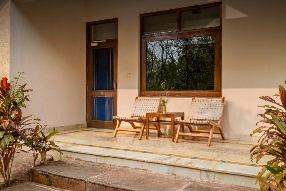 Porch seating area with two woven chairs and a small wooden table in front of a door and large window, framed by tropical plants on both sides.