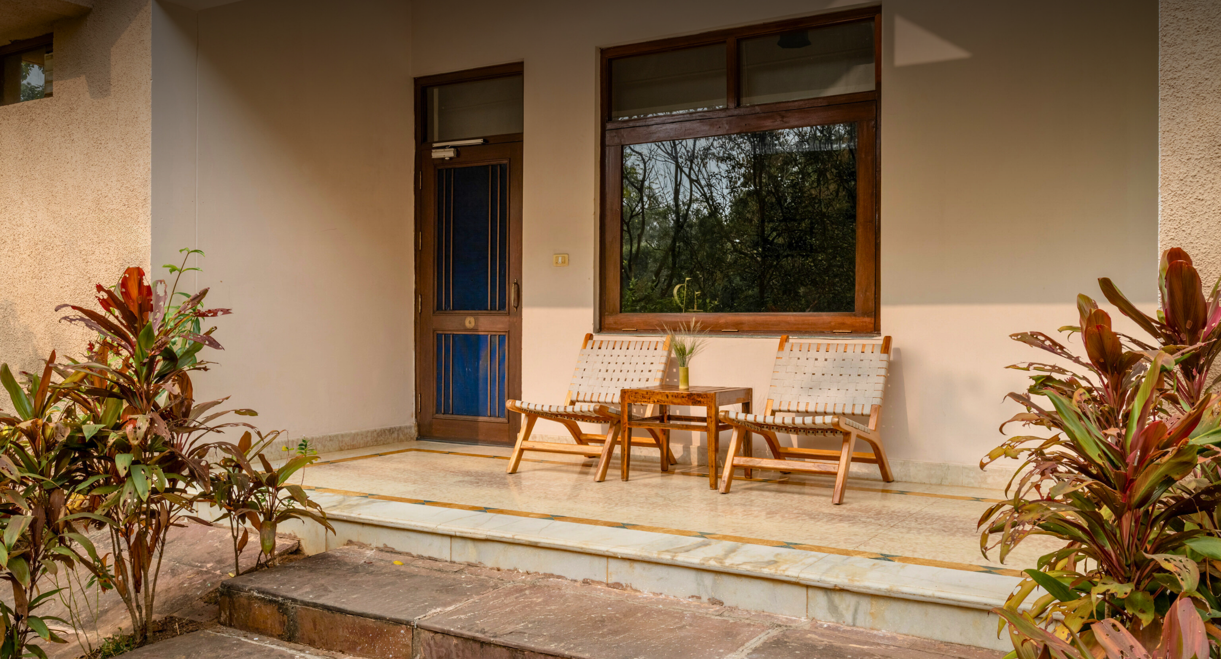 Front porch seating area with a brown door, large window, two woven chairs, and a small wooden table between them, bordered by tropical plants.
