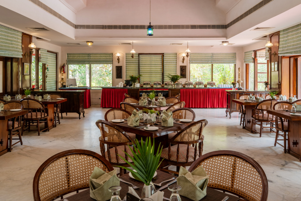 Dining hall with dark wood tables and woven chairs, folded napkins at each place, and a long buffet with red skirts along the back wall.