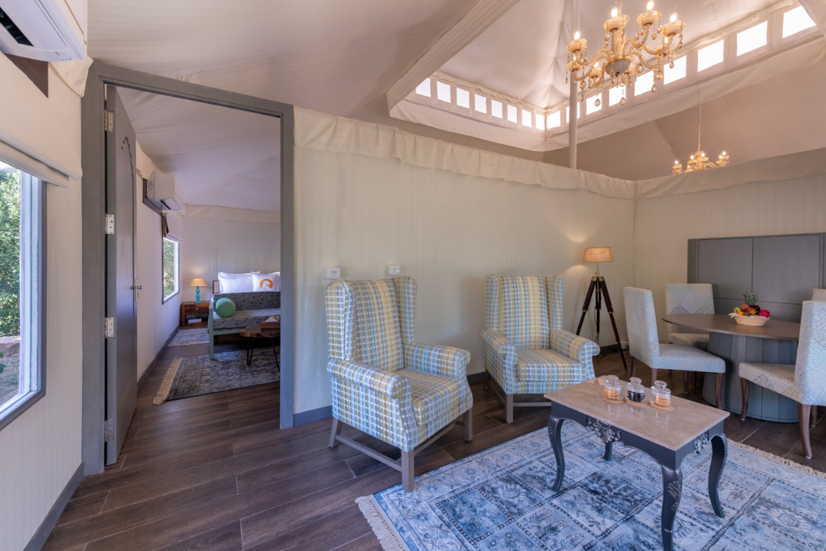 Living room: two plaid armchairs around a coffee table on a blue rug, with a dining nook and chandelier overhead; doorway to a bedroom in the back.