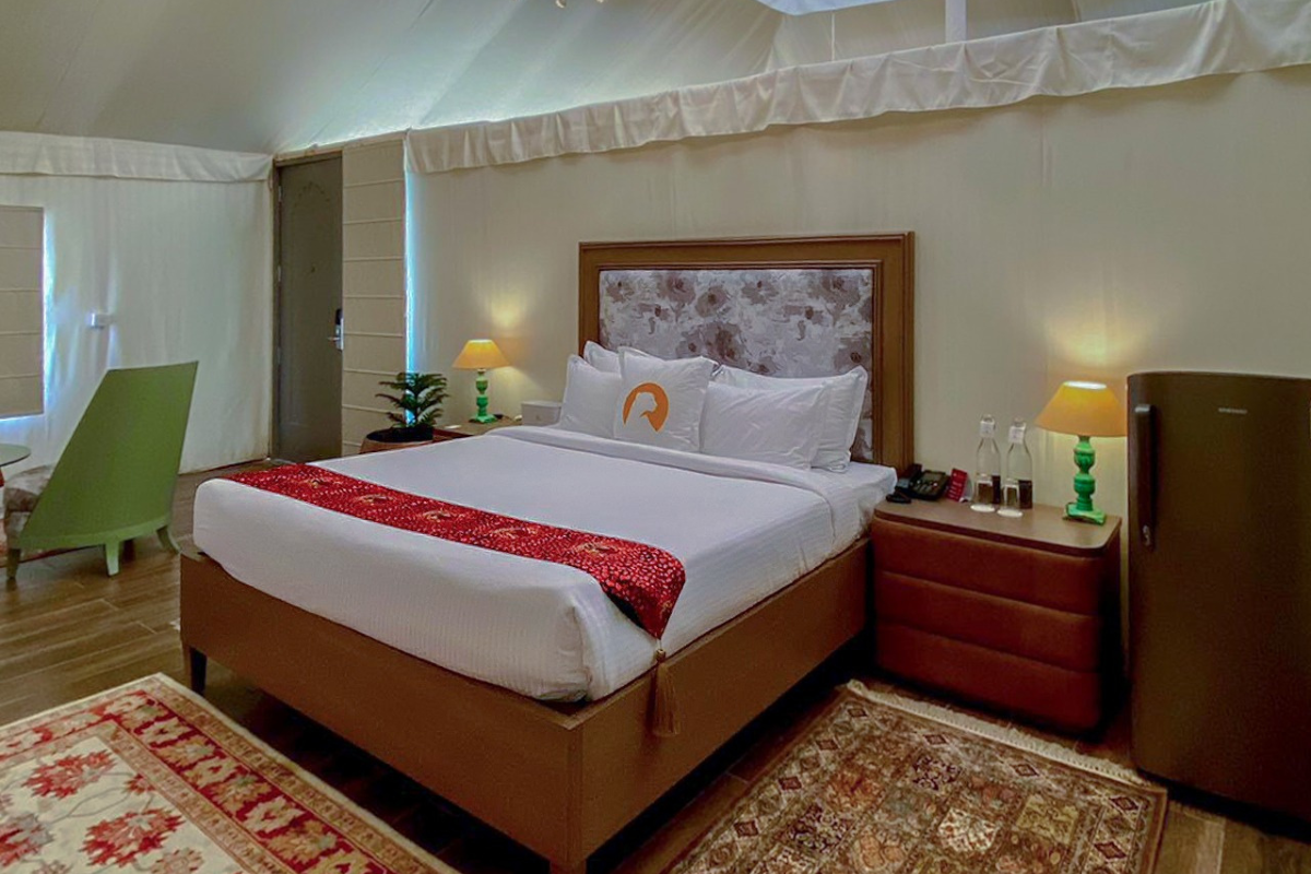 Hotel bedroom with a large bed, floral headboard, white linens, and a red decorative runner, flanked by nightstands and lamps—fridge and seating visible nearby