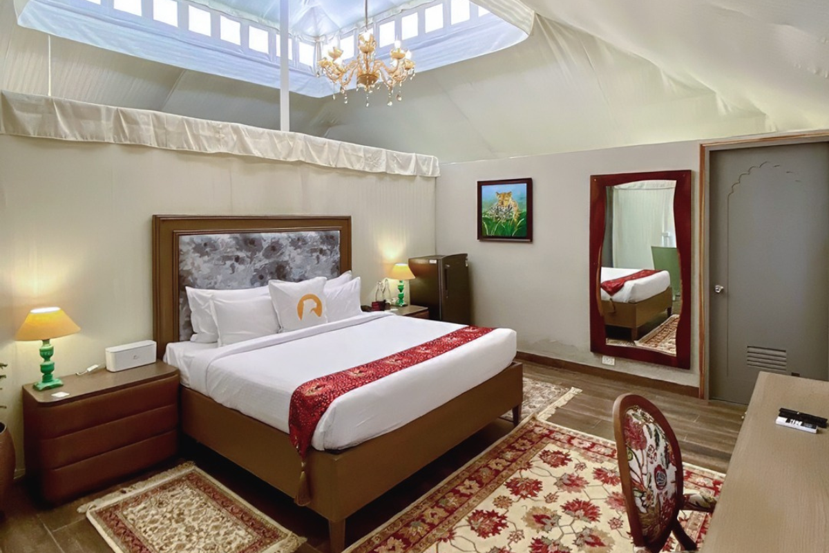 King bed with white linens and red bed runner, large padded headboard, two nightstands, lamps, and a crystal chandelier in a hotel room