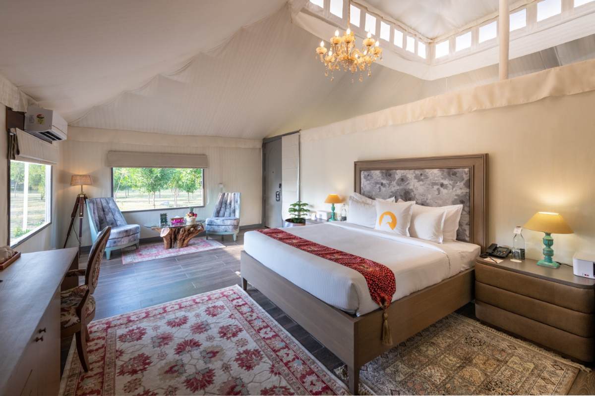 Hotel room inside a white tent with a chandelier, king bed, and a seating area by a window; several area rugs add color and texture.