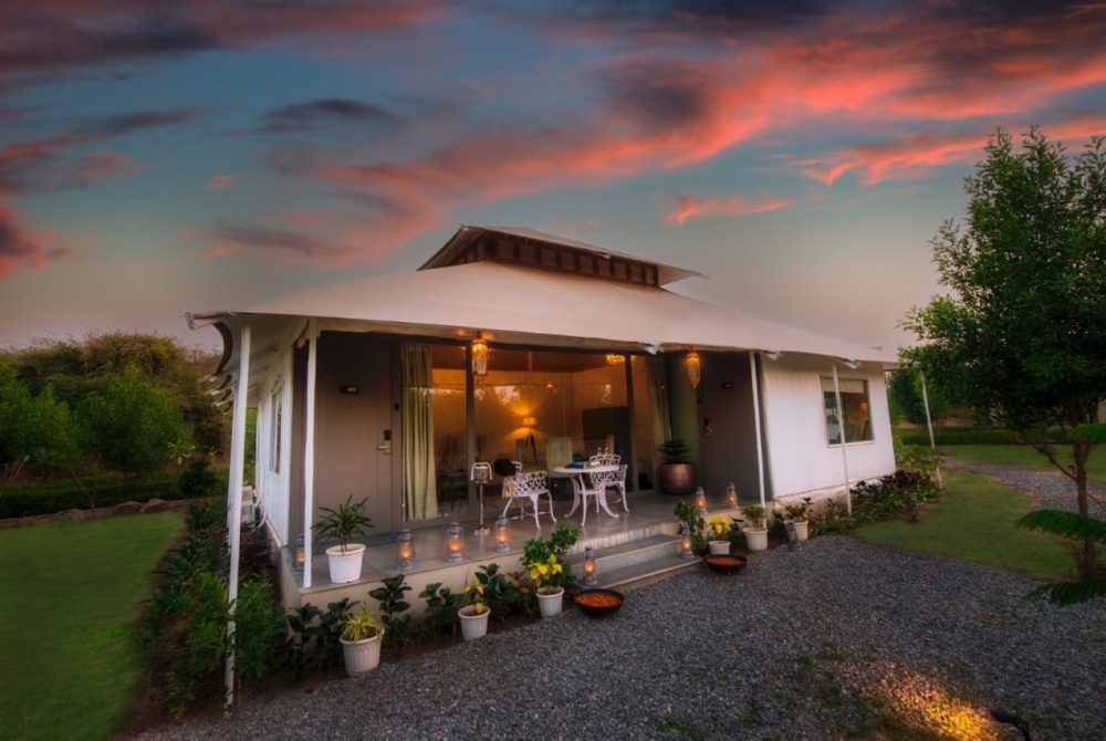 White single-story cottage with a covered porch, glass doors, and white metal seating, lit by lanterns at dusk. Potted plants line a gravel path.