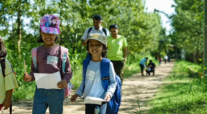 Two smiling children with backpacks walk along a sunny park trail, holding papers in their hands.