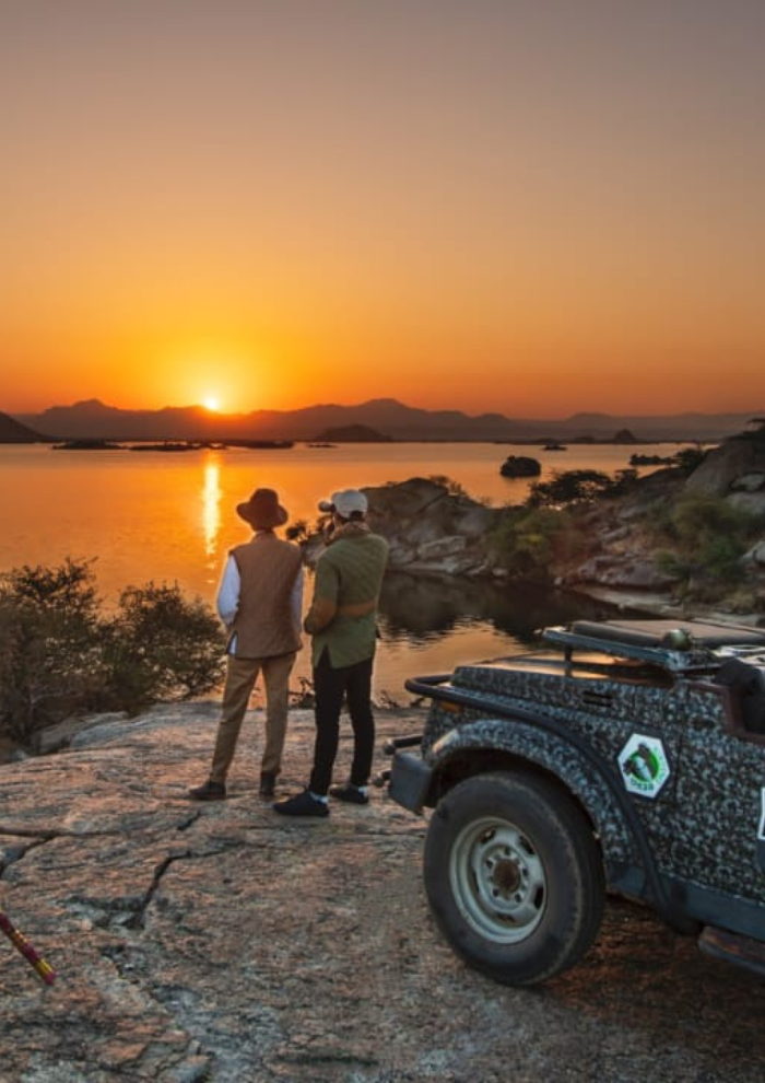 LAKESIDE BREAKFAST Two people wearing hats stand on a rocky lakeshore watching a orange sunset, with a camo-pattern off-road vehicle parked nearby.