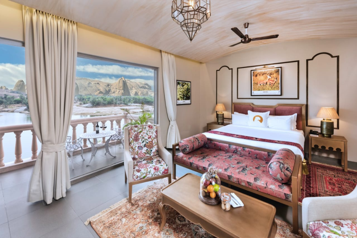 Luxurious hotel room with a king bed, floral bench, and a balcony overlooking a lake and rocky landscape outside.