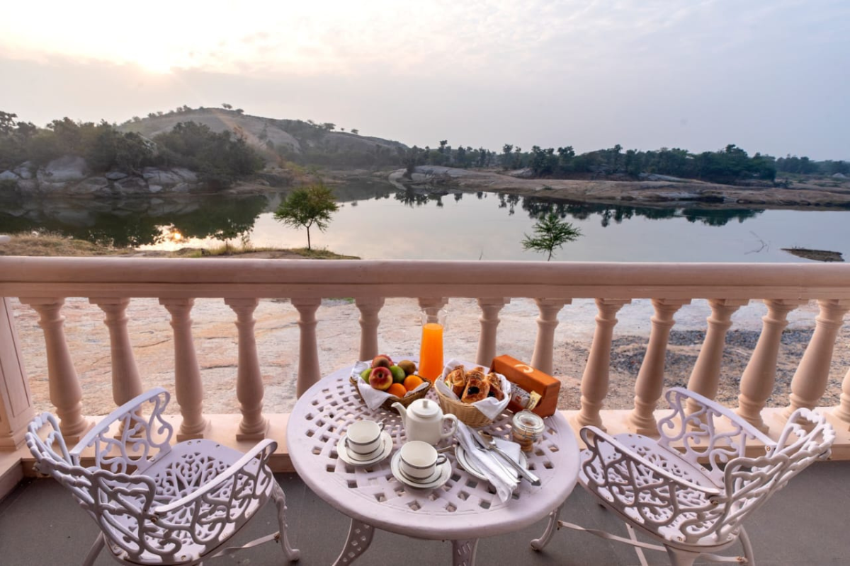 Breakfast on a white wrought-iron balcony table with fruit, croissants, teacups, and a candle, overlooking a calm lake and hills at sunrise.