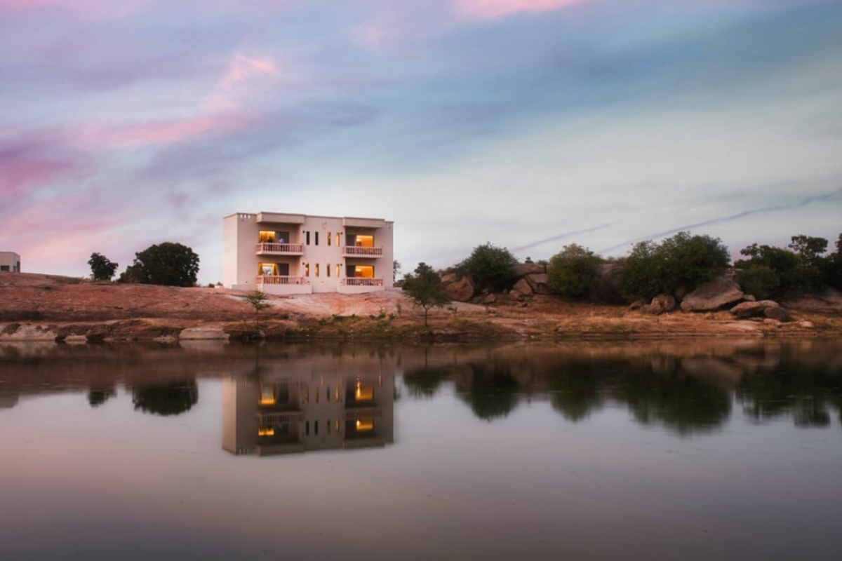 White two-story house on a rocky riverbank, reflected in calm water at sunset with pink and blue sky.