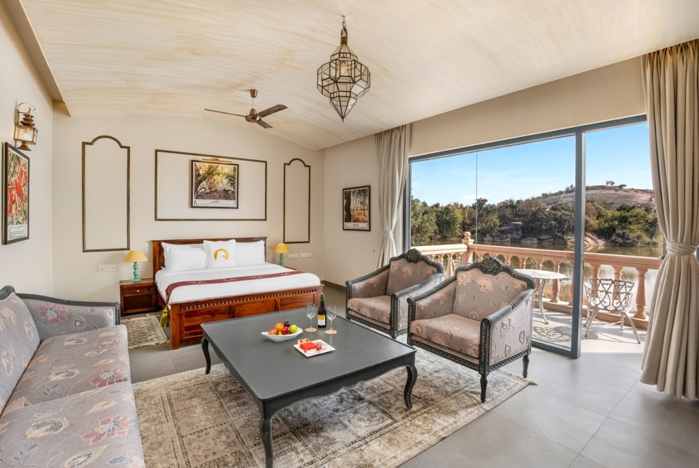 Luxury hotel suite: wood bed, two upholstered chairs, coffee table with fruit and wine, and a balcony overlooking a river and hillside landscape.